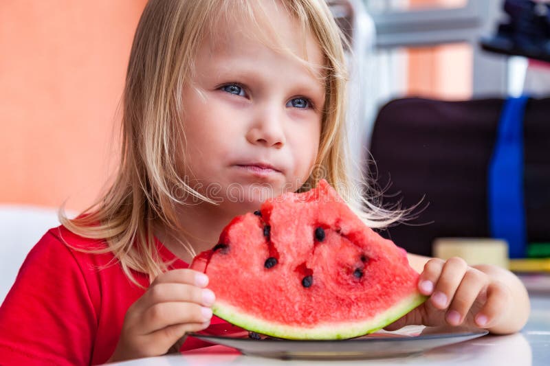 Lustigt, blond barnporträtt med vattenmelon inomhus som ser bort. Söt liten småflicka 4 år som äter vattenmelon royaltyfri foto