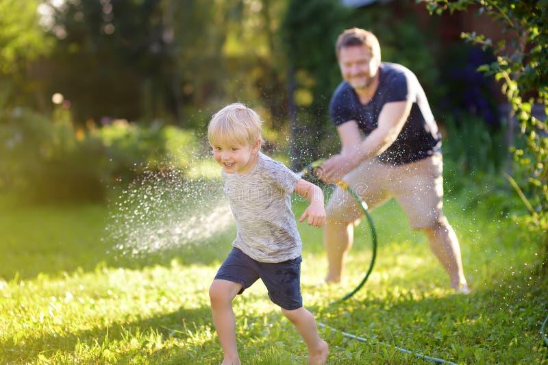 Lustigt liten pojke med sin pappa som leker med trädgårdsslang på solig bakgård. Barn som har kul med vattensprejning royaltyfria foton