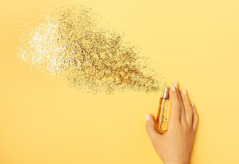 Luxury perfume concept. Female hand holding stylish bottle of perfume with spray of sparkles on yellow background