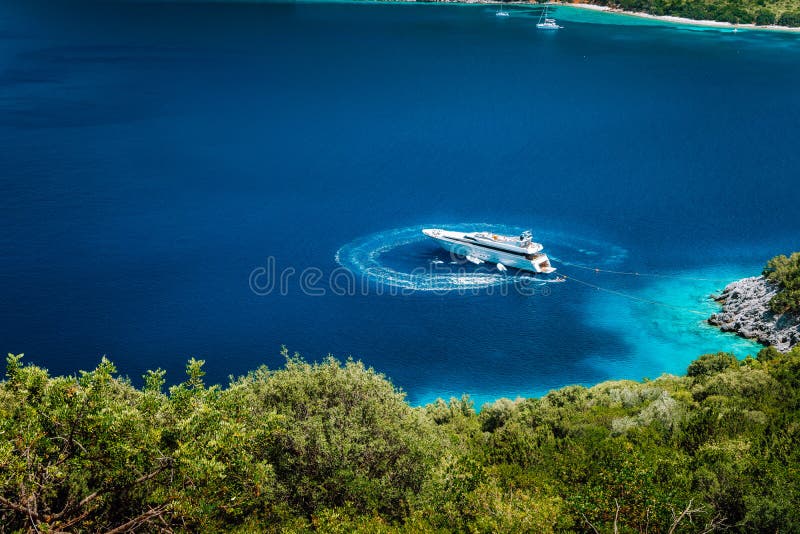 Luxury white yacht sail boat anchoring in a tranquil bay in deep blue water water, near picturesque shore of greek