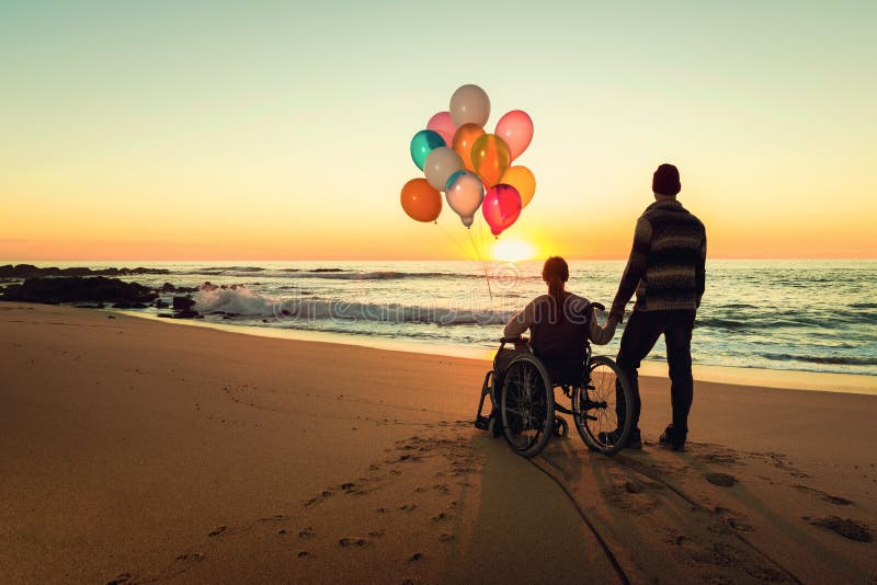 Happy couple at the beach, where women is on a wheelchair holding balloons on her hands. Happy couple at the beach, where women is on a wheelchair holding balloons on her hands
