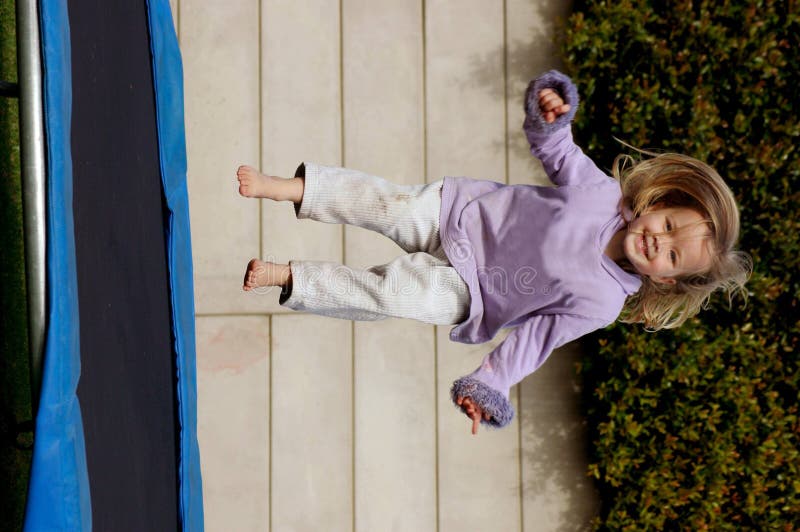 A full body of a beautiful little caucasian white girl child with blond long hair and happy cute expression in her pretty face having great fun by jumping on a trampoline in the backyard of a playschool outdoors. A full body of a beautiful little caucasian white girl child with blond long hair and happy cute expression in her pretty face having great fun by jumping on a trampoline in the backyard of a playschool outdoors