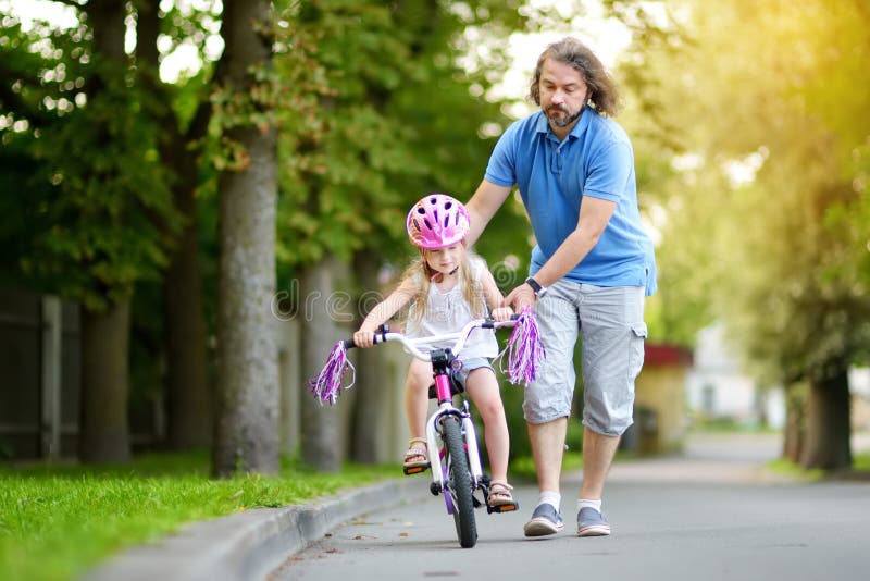 Lycklig fader som undervisar hans lilla dotter att rida en cykel Barn som lär att rida en cykel royaltyfria bilder