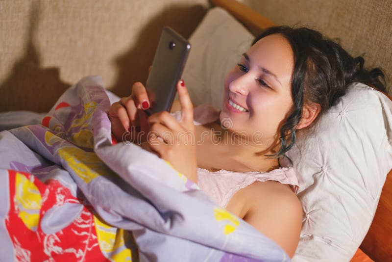 Lycklig le ung kvinna som smsar på smartphonen i hemmastatt sovrum för säng på natten arkivbild Lycklig le ung kvinna som smsar på smartphonen i hemmastatt sovrum för säng på natten arkivbild