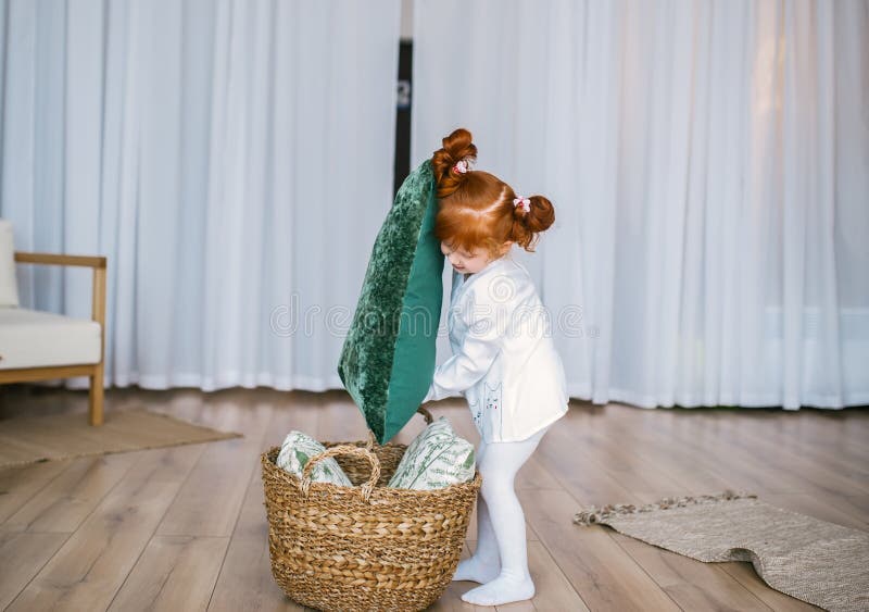 Lycklig rödhårig manliten flicka i pyjamaslek med korgen och kuddar på golv hemma royaltyfri bild Lycklig rödhårig manliten flicka i pyjamaslek med korgen och kuddar på golv hemma royaltyfri bild