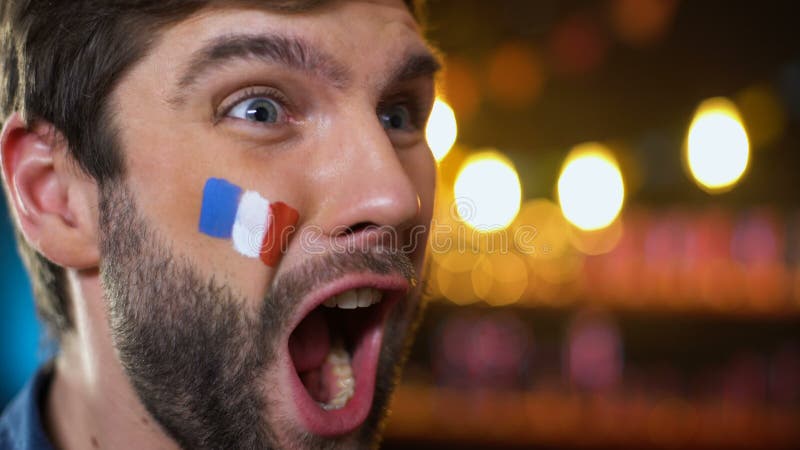 Lycklig stilig skäggig fransk fan med den målade flaggan på kind som firar mål