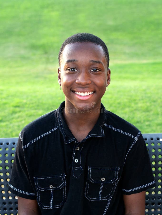 A happy, ,African American, teen boy smiles while sitting on a bench outside. A happy, ,African American, teen boy smiles while sitting on a bench outside