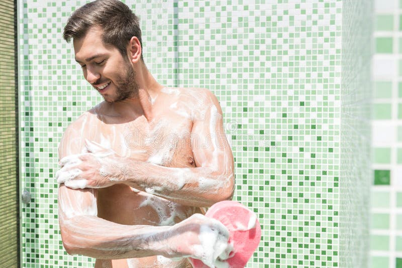 Cheery youthful man with beard is washing body with soap in shower unit. He is holding puff and looking at his shoulder with smile. Focus on chest. Cheery youthful man with beard is washing body with soap in shower unit. He is holding puff and looking at his shoulder with smile. Focus on chest