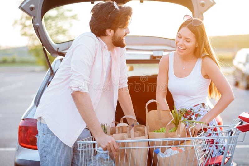 Lyckliga unga par som går tillbaka från livsmedelsbutikshopping som laddar pappers- påsar med mat in i en bilstam arkivbilder Lyckliga unga par som går tillbaka från livsmedelsbutikshopping som laddar pappers- påsar med mat in i en bilstam arkivbilder
