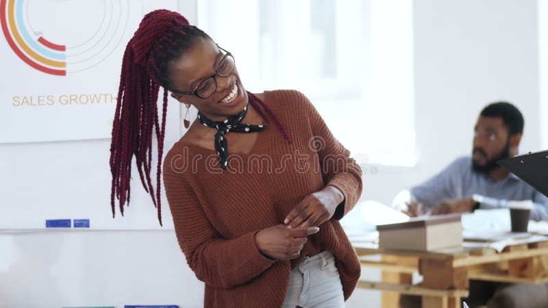 Lyckligt ungt tillfälligt afrikanskt kvinnaframstickande i den ledande diskussionen för glasögon som pratar till kollegor på det