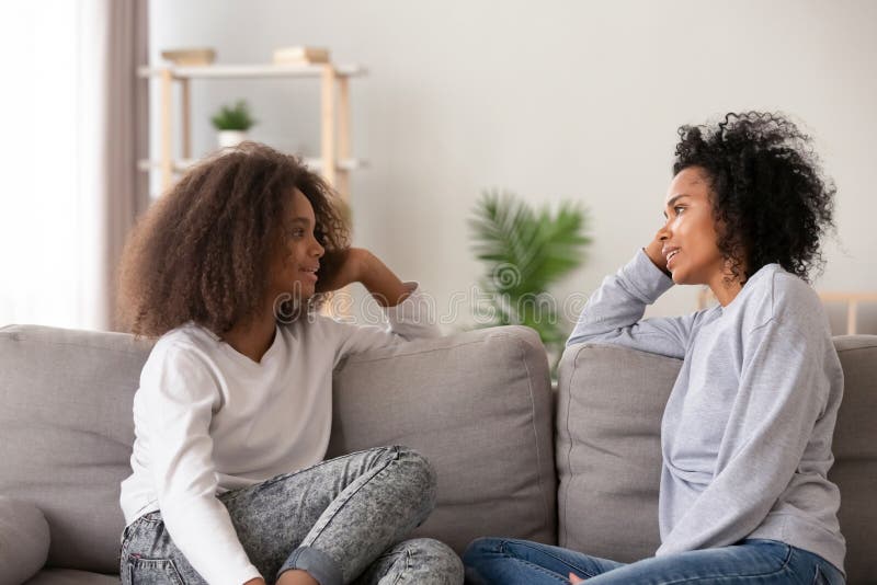 Young african black mother chatting with teen daughter, older sister or friendly nanny and adolescent girl talking sitting together on sofa at home. Good relations different aged people trust concept. Young african black mother chatting with teen daughter, older sister or friendly nanny and adolescent girl talking sitting together on sofa at home. Good relations different aged people trust concept