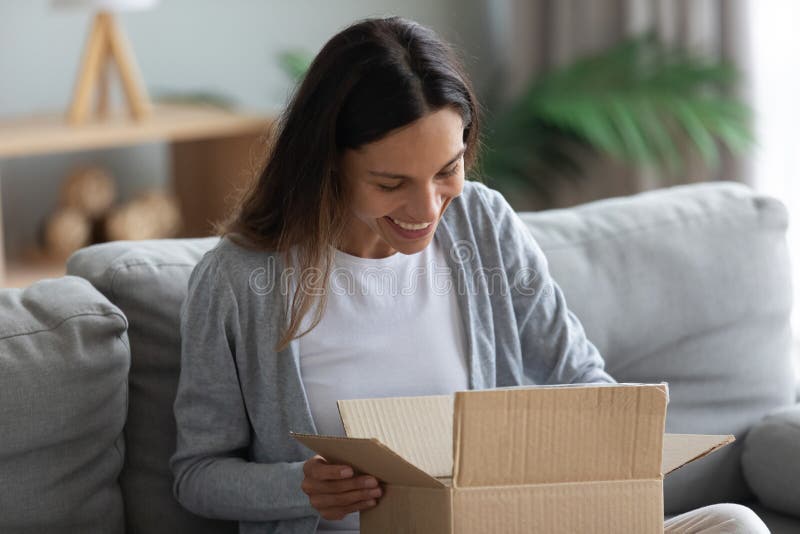 Smiling young woman unpacking parcel sitting on couch at home, pretty curious girl opening cardboard box close up, satisfied client received online store order, good delivery service concept. Smiling young woman unpacking parcel sitting on couch at home, pretty curious girl opening cardboard box close up, satisfied client received online store order, good delivery service concept
