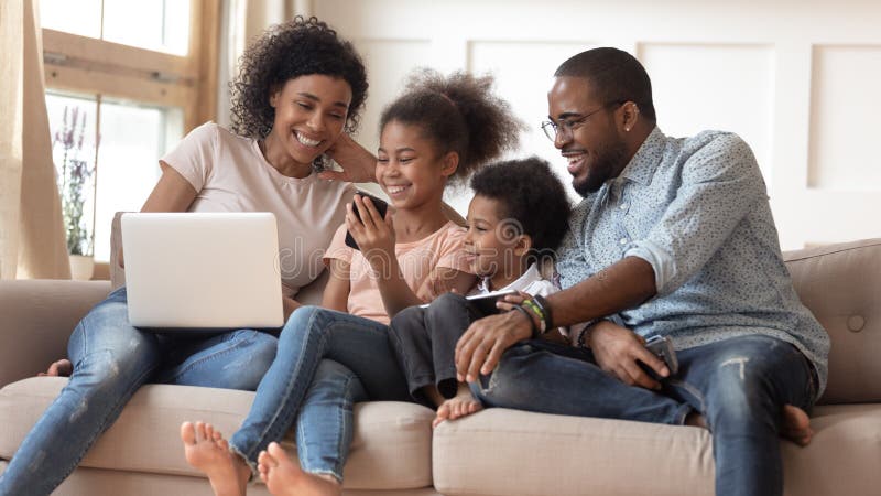 Happy african American young parents sit relax on couch with small kids watch funny video together, smiling black family with children rest on sofa with gadgets laugh enjoy cartoon on cellphone. Happy african American young parents sit relax on couch with small kids watch funny video together, smiling black family with children rest on sofa with gadgets laugh enjoy cartoon on cellphone