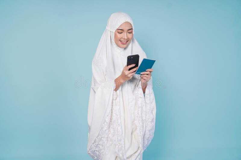 Lächelnde asiatische Muslimfrau im Gebetskleid Blick auf den Pass und Smartphone hält sie isoliert durch blauen Hintergrund. Hadsc stockbild