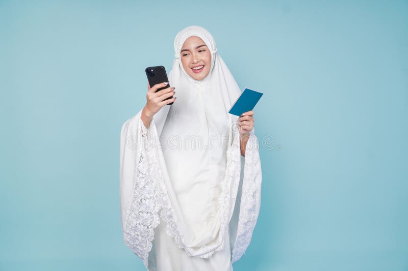 Lächelnde asiatische Muslimfrau im Gebetskleid Blick auf den Pass und Smartphone hält sie isoliert durch blauen Hintergrund. Hadsc lizenzfreies stockfoto