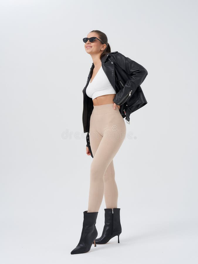 Lächelnde Frau in einer Sonnenbrille weiße Ernte Top beige Leggings und schwarze hochhackige Stiefel, die selbstbewusst mit Händen stockfotografie