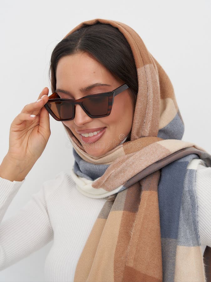 Lächelnde Frau mit dunkler Sonnenbrille, braun und blau gemusterter Schal, bedeckt ihren Kopf und ein weißes Stricktop. Ihre Linke lizenzfreies stockfoto