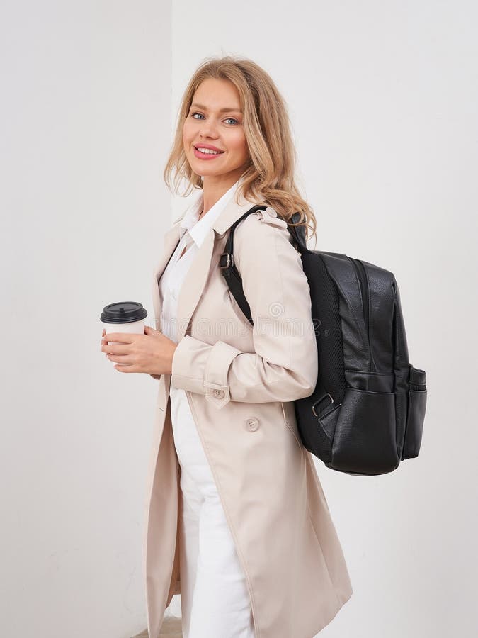 Lächelnde Frau mit langen Haaren mit beigem Mantel und weißem Hemd trägt einen schwarzen Rucksack und eine togo Kaffeetasse. Sie lizenzfreie stockfotografie