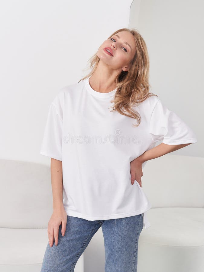 Lächelnde Frau mit langen Haaren, die ein weißes übergroßes Hemd trägt, und blaue Jeans stehen selbstbewusst mit Hand auf Hüfte stockfotografie