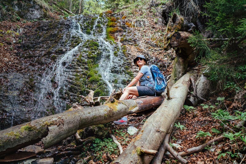 Lächelnde Frau mit Rucksack in einem Hut gekleidet in aktiven Trekkingbekleidung und Stiefeln sitzen in der Nähe des Bergflusses W lizenzfreie stockfotografie