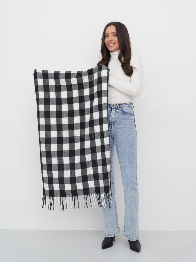 Lächelnde Frau in weißem Rollkragen und blauer Jeans hält eine große schwarz-weiß karierte Decke mit Rändern. Sie lizenzfreies stockbild