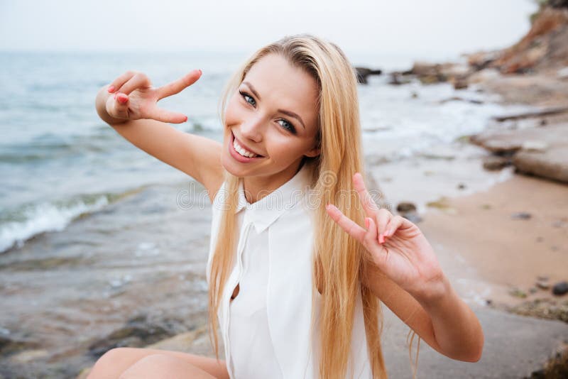 Lächelnde junge Blondine, die v-Zeichen am felsigen Strand zeigen stockfotografie Lächelnde junge Blondine, die v-Zeichen am felsigen Strand zeigen stockfotografie