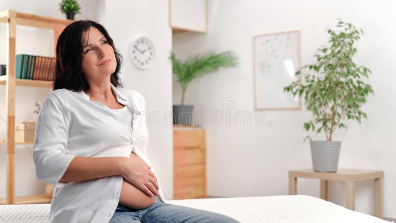 Lächelnde schöne schwangere Frau, die Bauch streichelt und mit positiver Emotion in hellweißem Raum träumt