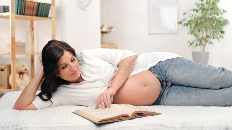 Lächelnde schöne schwangere Frau lesen Papierbuch entspannt liegen auf dem Bett in gemütlichen hellweißen Raum