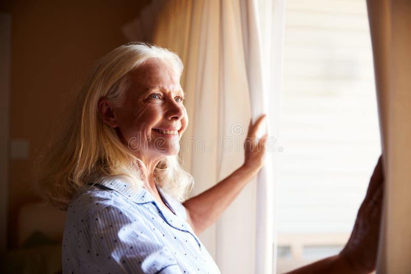 Lächelnde ältere weiße Frau, welche die Vorhänge auf einem sonnigen Morgen, Seitenansicht, Abschluss eröffnet lizenzfreie stockfotografie Lächelnde ältere weiße Frau, welche die Vorhänge auf einem sonnigen Morgen, Seitenansicht, Abschluss eröffnet lizenzfreie stockfotografie