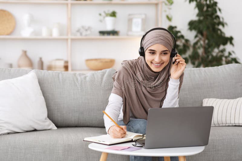 Lächelnder Arabisch Mädchen im Kopftuch und Kopfhörer, die zu Hause mit Notebook studieren