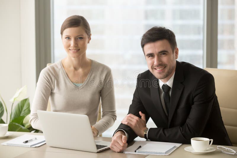 Smiling businessman and businesswoman looking at camera, confident successful boss and professional assistant posing at workplace sit in front of laptop, talented motivated business team portrait. Smiling businessman and businesswoman looking at camera, confident successful boss and professional assistant posing at workplace sit in front of laptop, talented motivated business team portrait