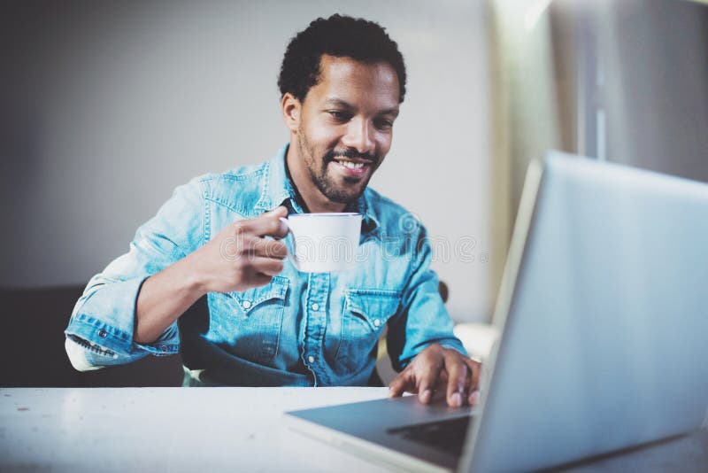 Lächelnder junger afrikanischer Mann, der Videogespräch über Laptop mit Partnern beim schwarzen Tee der weißen Schale herein trin stockbild Lächelnder junger afrikanischer Mann, der Videogespräch über Laptop mit Partnern beim schwarzen Tee der weißen Schale herein trin stockbild