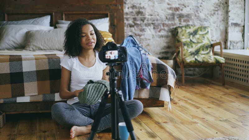 Lächelndes Videoblog der gelockten Afroamerikanermädchen-Aufnahme über ihre Garderobe für Reise mit dslr Kamera zu Hause