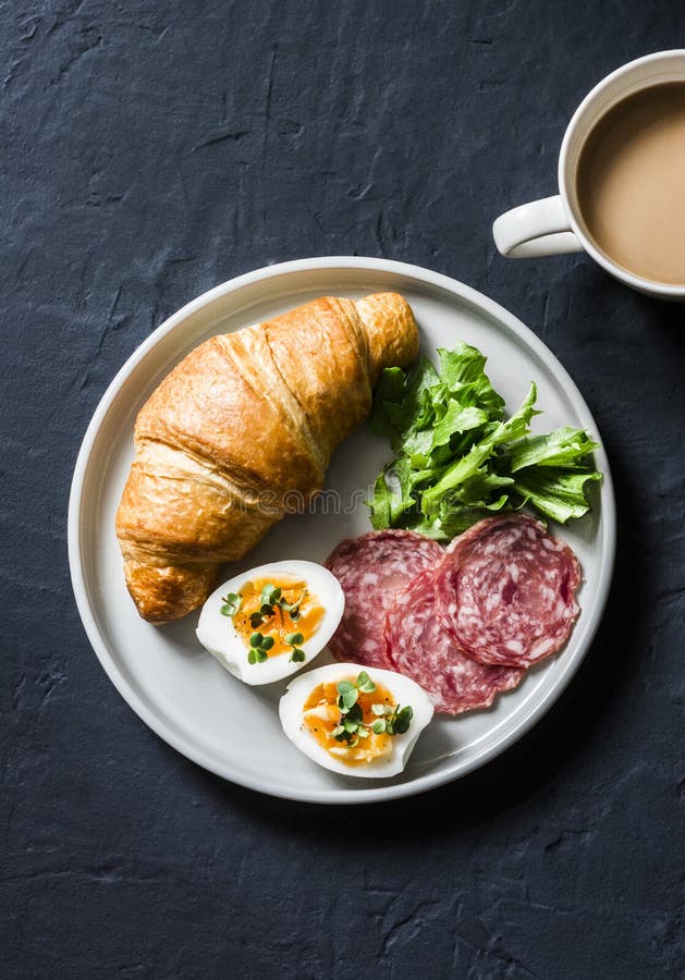 Läcker frunch eller frukost - gifflet, det kokta ägget, salamikorven, grön sallad och kaffe med mjölkar på en mörk bakgrund royaltyfri foto Läcker frunch eller frukost - gifflet, det kokta ägget, salamikorven, grön sallad och kaffe med mjölkar på en mörk bakgrund royaltyfri foto