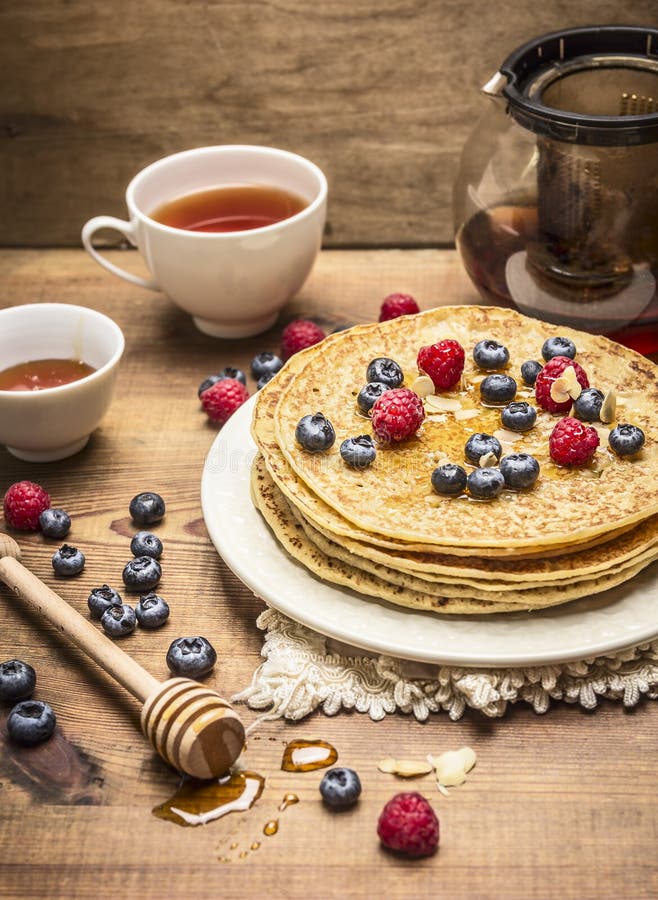 Läckra pannkakor med nya bär på en vit platta med den honungskeden och kopp te på träbakgrundsslut upp royaltyfria bilder Läckra pannkakor med nya bär på en vit platta med den honungskeden och kopp te på träbakgrundsslut upp royaltyfria bilder