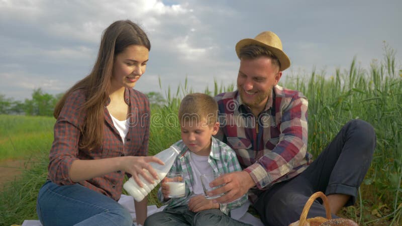 Ländliche Familie, auslaufende Milch der glücklichen Frau in Glas zum Sohn und Ehemann während des Picknicks in der Natur auf dem