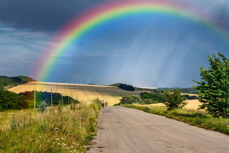 Ländliche Landschaft des Sommers - Straße mit Radfahrern in den Strahlen der untergehenden Sonne auf entferntem Regenbogen des Hi lizenzfreie stockfotos Ländliche Landschaft des Sommers - Straße mit Radfahrern in den Strahlen der untergehenden Sonne auf entferntem Regenbogen des Hi lizenzfreie stockfotos