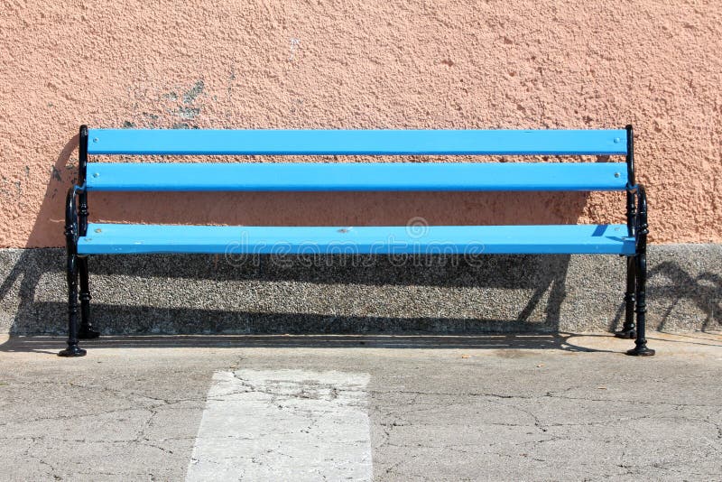Lång blå träoffentlig bänk med den svarta järnramen som framme monteras på sida av den stenlade trottoaren av husväggen royaltyfri fotografi Lång blå träoffentlig bänk med den svarta järnramen som framme monteras på sida av den stenlade trottoaren av husväggen royaltyfri fotografi