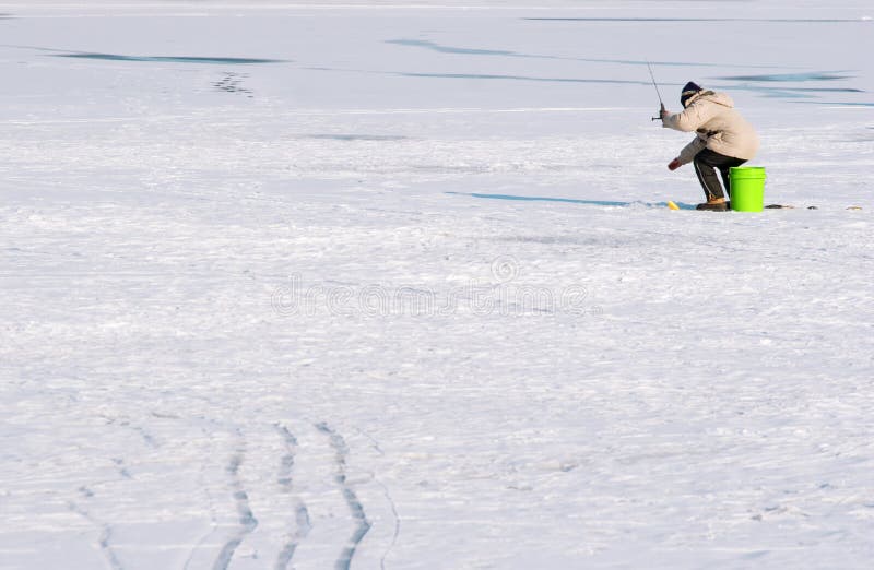 A person sits on a bucket on a frozen lake with a fishing pole. A person sits on a bucket on a frozen lake with a fishing pole.