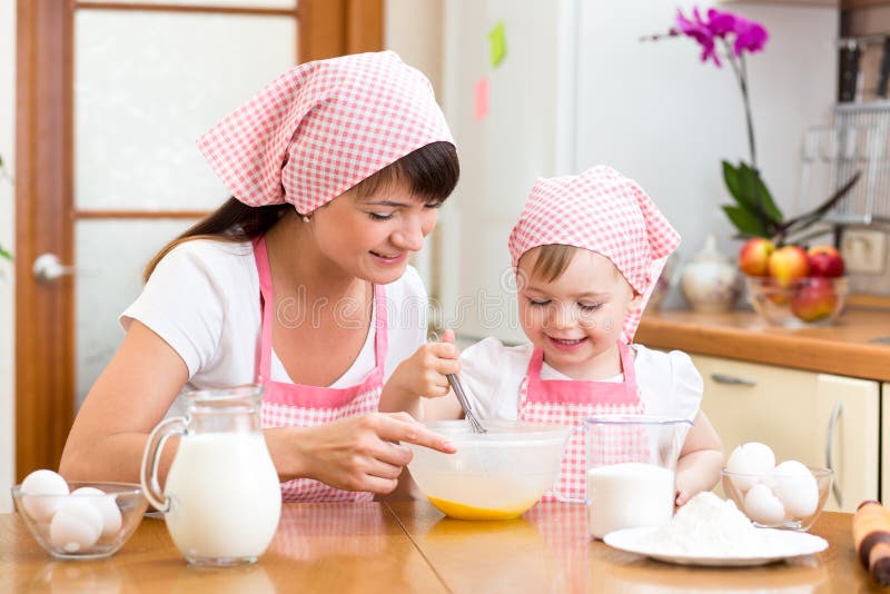 heureuse mère et fille cuisent ensemble dans la cuisine, utilisent  différents ingrédients, portent des tabliers, se tiennent contre  l'intérieur de la cuisine, la fille verse du lait à l'arc. maman  attentionnée apprend