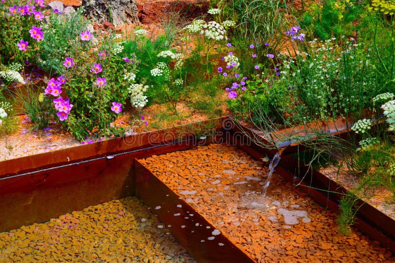 The M & G Show Garden at the RHS Chelsea Flower Show in London May 2018. Designed by Sarah Price it shows hard landscaping made of packed mud with water fountains and pools plus grass and highlight coloured flowers. The M & G Show Garden at the RHS Chelsea Flower Show in London May 2018. Designed by Sarah Price it shows hard landscaping made of packed mud with water fountains and pools plus grass and highlight coloured flowers.