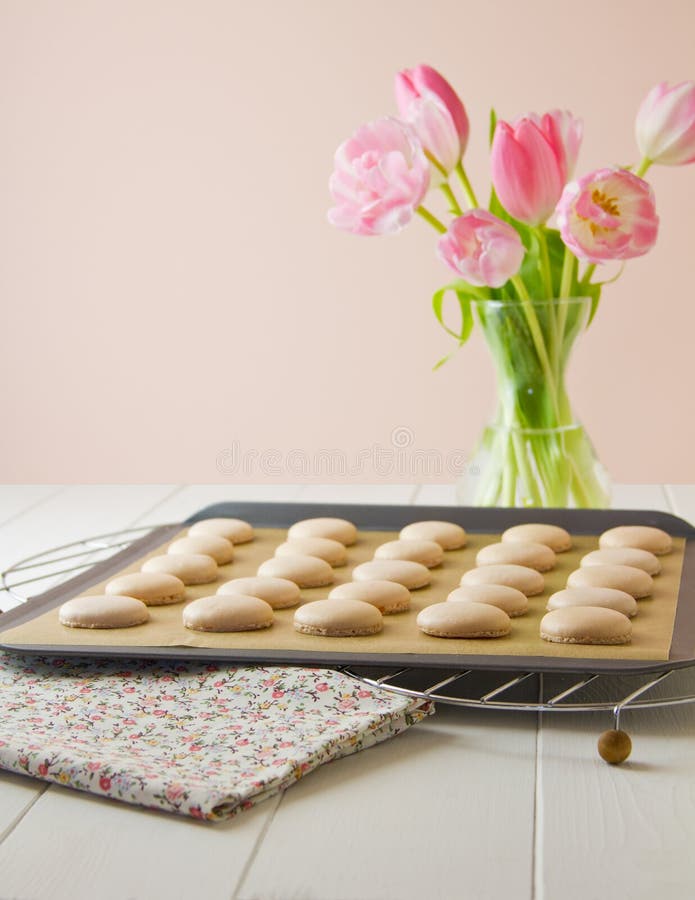 25 French macaron shells (pronounced macaroon, a popular buttercream filled meringue type cookie or biscuit) on baking sheet lined with baking parchment paper, on wire cooling racks. On white wood table with floral tea towel. Soft pink and white tulips sit to one side. 25 French macaron shells (pronounced macaroon, a popular buttercream filled meringue type cookie or biscuit) on baking sheet lined with baking parchment paper, on wire cooling racks. On white wood table with floral tea towel. Soft pink and white tulips sit to one side.