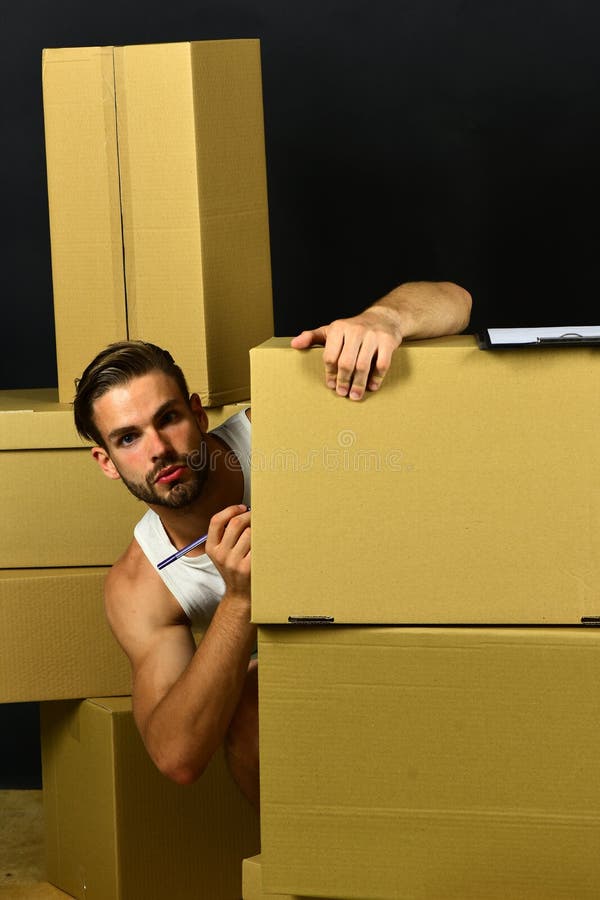 Macho with beard and distracted face. Man with strong arms labeling cardboard boxes. Guy with clip board and pen isolated on black background. Delivery and moving in concept. Macho with beard and distracted face. Man with strong arms labeling cardboard boxes. Guy with clip board and pen isolated on black background. Delivery and moving in concept.