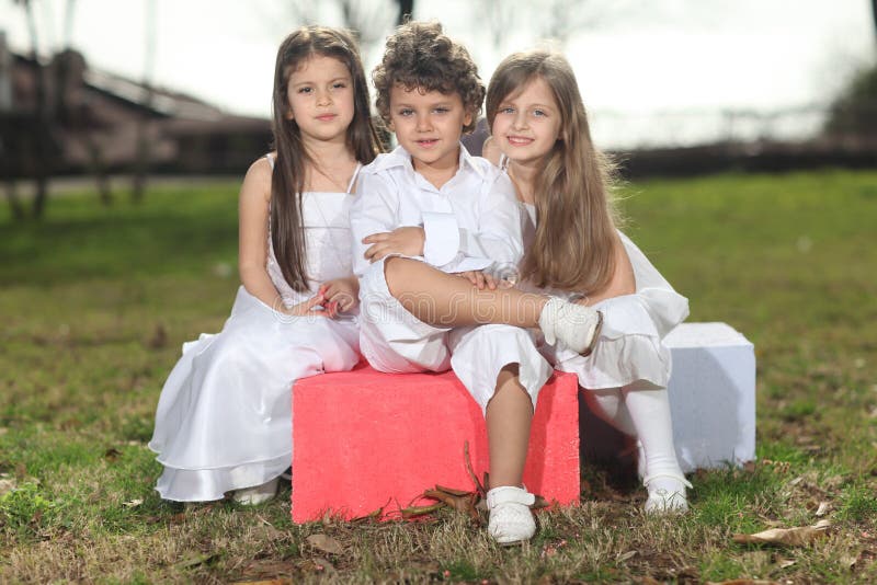 Cute young boy with two beautiful young blonde girls with white dresses sitting on colorful cubes, smiling to the camera, sun reflection in hair, backlight. Cute young boy with two beautiful young blonde girls with white dresses sitting on colorful cubes, smiling to the camera, sun reflection in hair, backlight