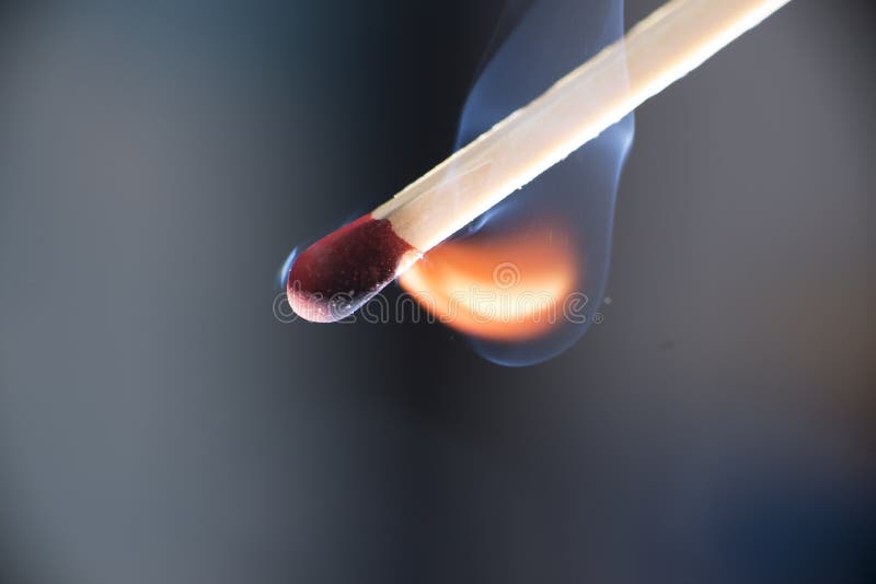 Macro photo of match at moment of ignition, red tip still intact, orange flame starting underneath, blue smoke. Isolated on blue-gray background. Macro photo of match at moment of ignition, red tip still intact, orange flame starting underneath, blue smoke. Isolated on blue-gray background.