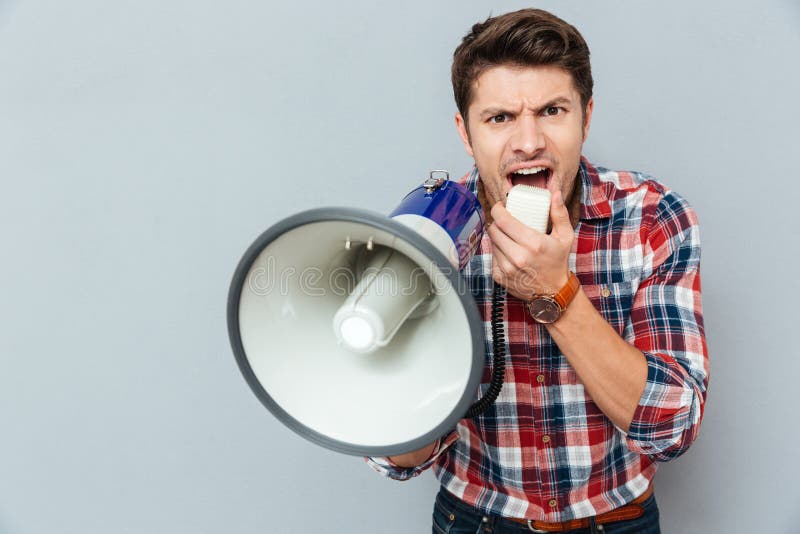 Mad crazy young man in plaid short shouting in megaphone over grey background