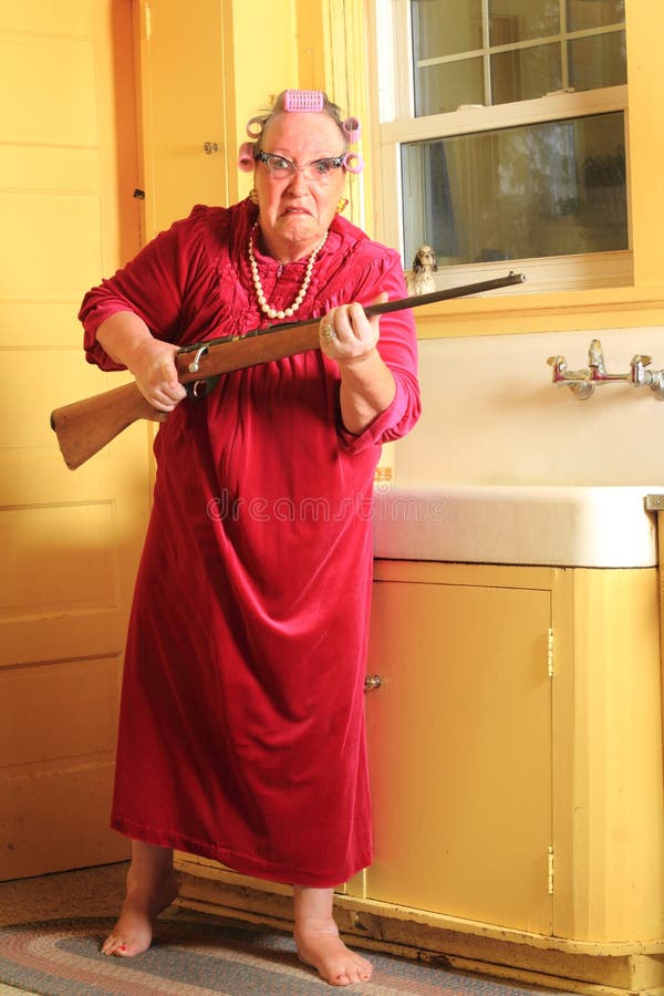 Mad Granny with Rifle. A mad senior gray haired granny lady wearing cat eye glasses, pearls and curlers in her hair in an old fashioned yellow kitchen standing Mad Granny with Rifle. A mad senior gray haired granny lady wearing cat eye glasses, pearls and curlers in her hair in an old fashioned yellow kitchen standing