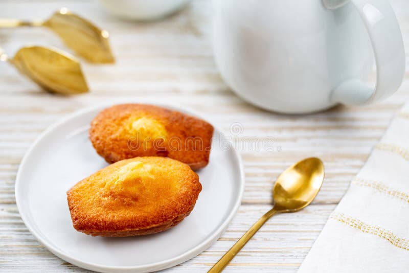 Madeleine cookie or French sponge cake in a white plate alongside white teapot, golden spoon and tong by a white napkin royalty free stock photo Madeleine cookie or French sponge cake in a white plate alongside white teapot, golden spoon and tong by a white napkin royalty free stock photo