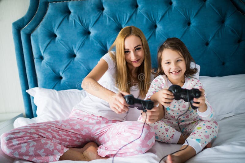 Madre alegre e hija que juegan a los videojuegos en cama en casa imagen de archivo Madre alegre e hija que juegan a los videojuegos en cama en casa imagen de archivo