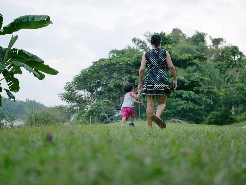 Madre asiática que mantiene la mano de su pequeña hija y tener un paseo unida en un campo de hierba verde al aire libre por la or fotos de archivo libres de regalías Madre asiática que mantiene la mano de su pequeña hija y tener un paseo unida en un campo de hierba verde al aire libre por la or fotos de archivo libres de regalías